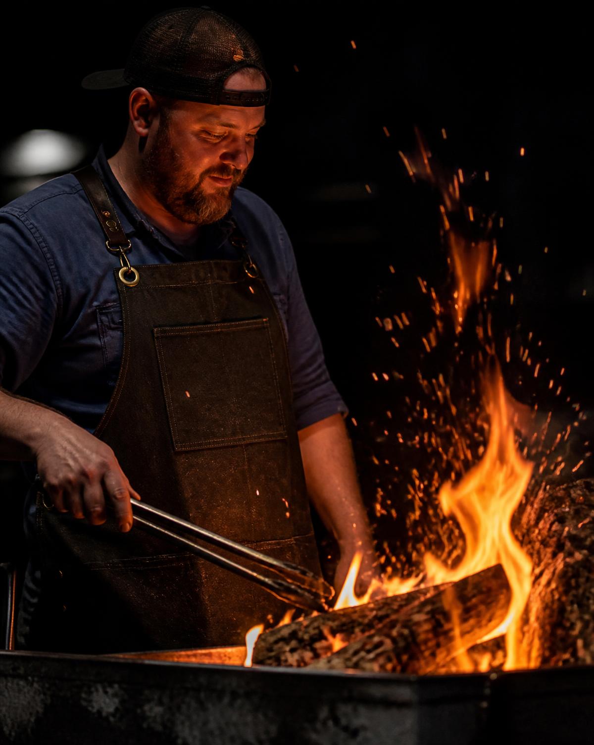 Cook tending glowing oak coals with long tongs beside a Santa Maria style grill